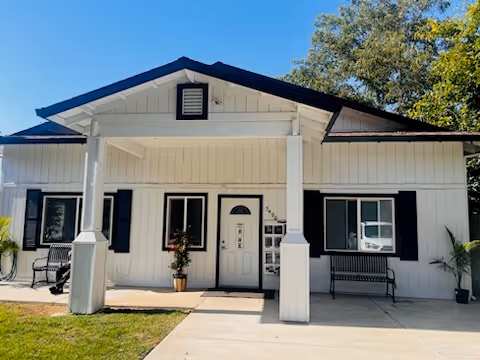 Front exterior view of a single-story white building with black trim and a covered porch supported by two columns. There are two windows with black shutters on either side of a central door. A bench and potted plants are placed on the porch. The building is surrounded by a small lawn and trees in the background.