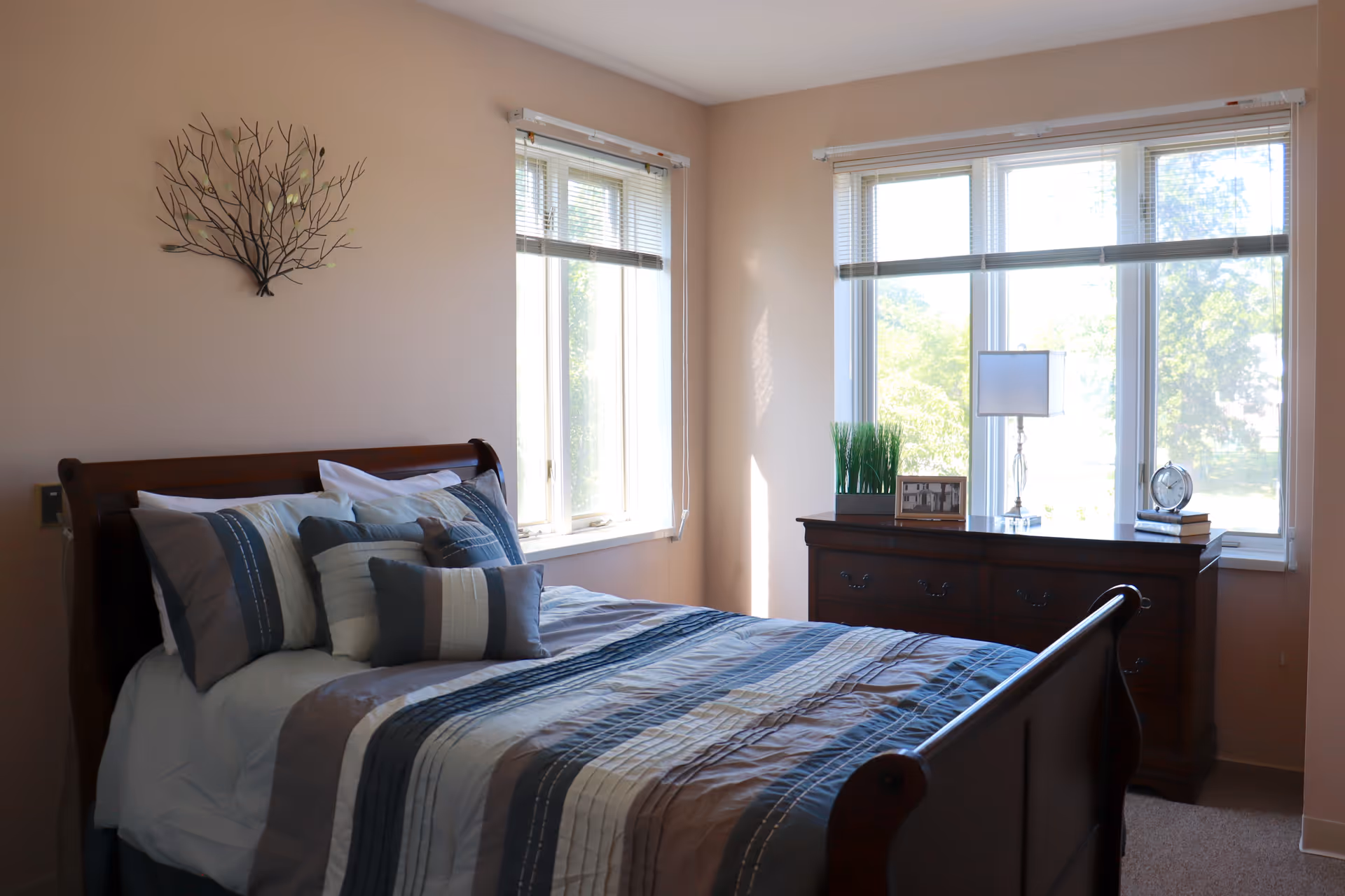 A cozy bedroom with a wooden bed frame and a neatly made bed featuring multiple pillows and a striped comforter in shades of blue, gray, and white. The room has beige walls, two windows with blinds letting in natural light, and a wooden dresser with decorative items including a lamp, a small plant, a clock, and framed photos.