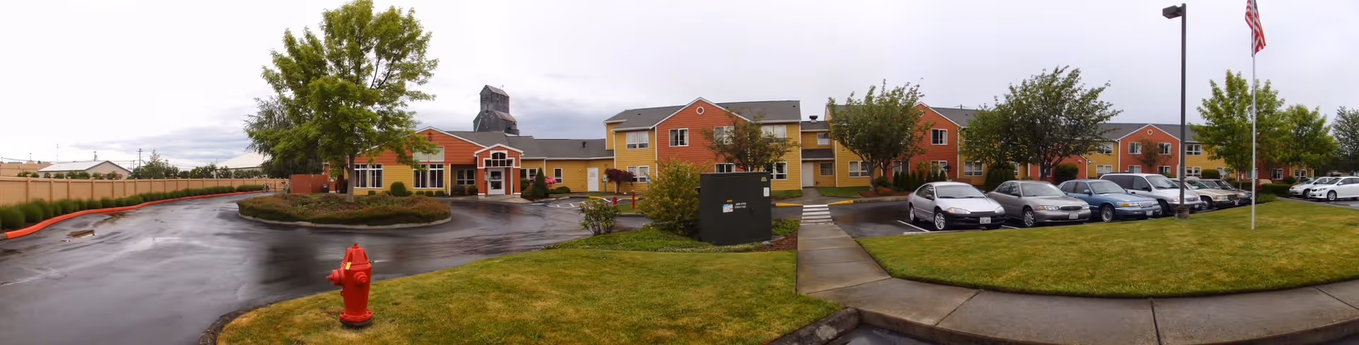 Panoramic exterior view of a two-story retirement community building with a driveway, parking lot, lawn, flagpole and a red fire hydrant in front.