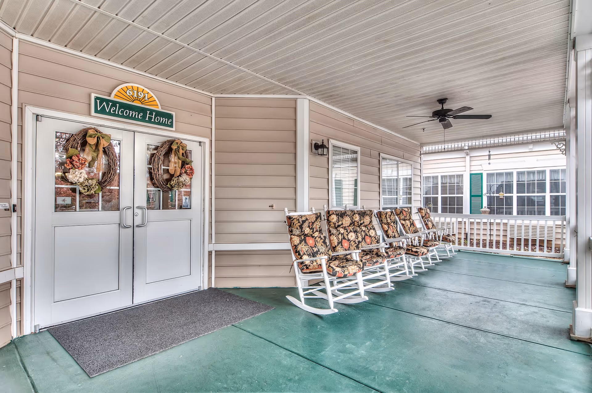 Covered porch area with six white rocking chairs featuring floral cushions lined up against the wall. Double doors with glass panels have two decorative wreaths and a sign above that reads 'Welcome Home'. The porch has a green floor and a ceiling fan, with windows on the adjacent wall.