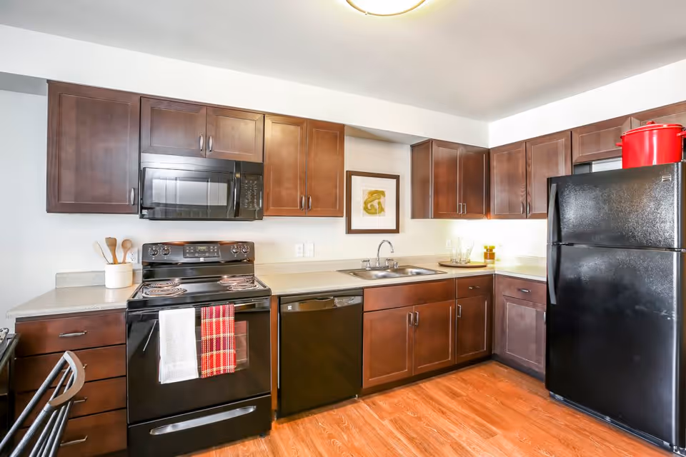 Modern kitchen with dark wooden cabinets, black appliances including a stove, microwave, dishwasher, and refrigerator. The kitchen has a double sink, light-colored countertops, wooden flooring, and a framed artwork on the wall.