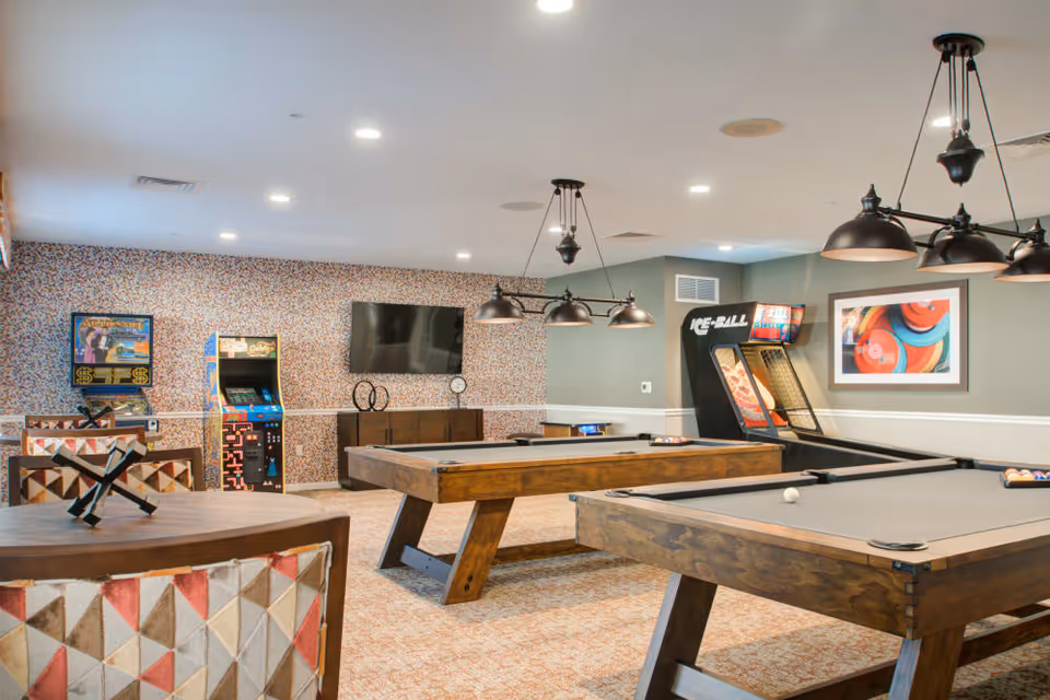 A recreational room with two wooden pool tables, arcade game machines, a wall-mounted flat screen TV, and modern pendant lighting. The room has patterned carpet and wallpaper, with a framed colorful abstract artwork on the wall.