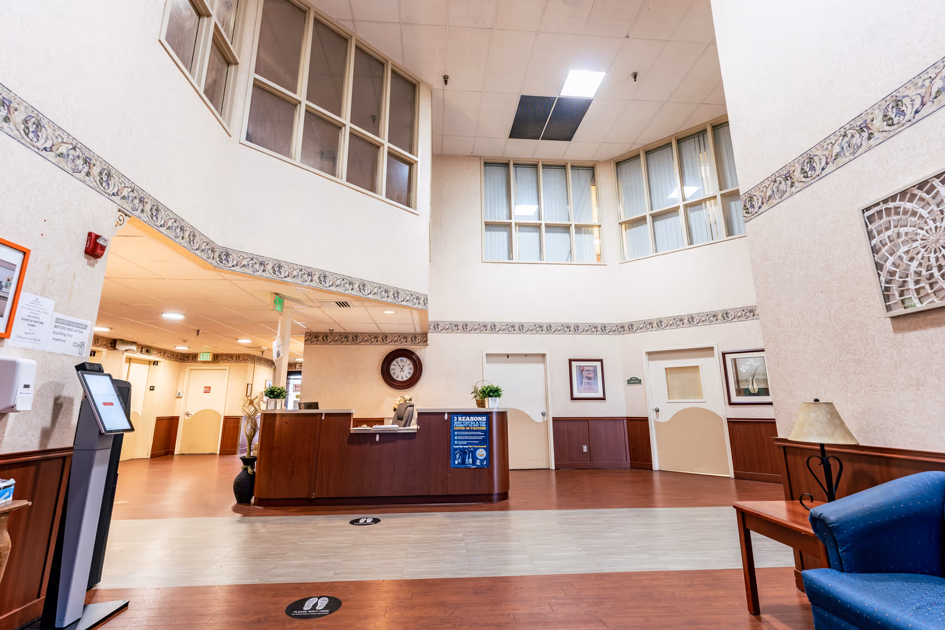 Interior view of a healthcare facility reception area with a wooden front desk, a clock on the wall, decorative wall trim, and several doors leading to other rooms. There is a blue armchair and a small wooden table with a lamp on the right side. The ceiling is high with large windows above the doors.
