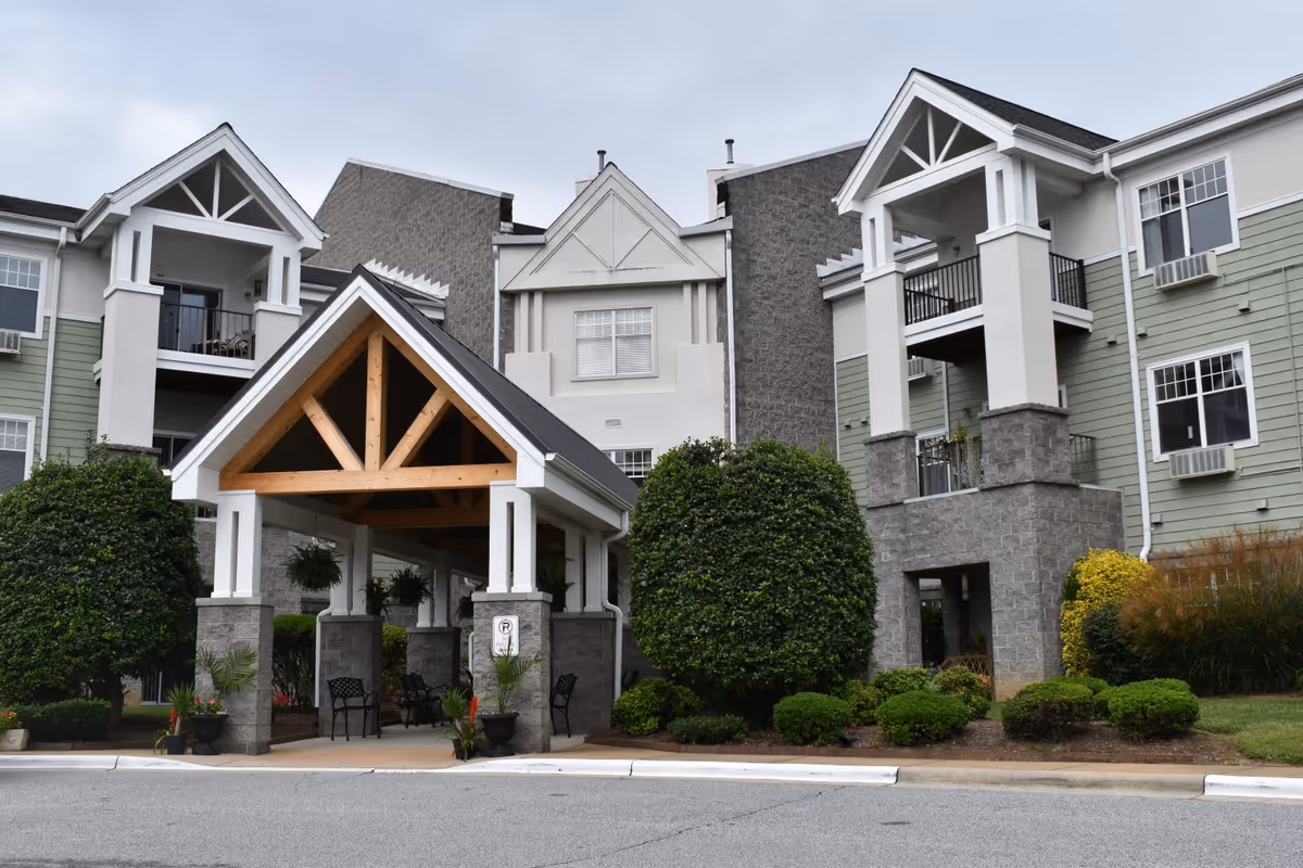 Front exterior view of Trinity View Retirement Community building with a covered entrance supported by stone pillars and wooden beams, surrounded by neatly trimmed bushes and greenery.