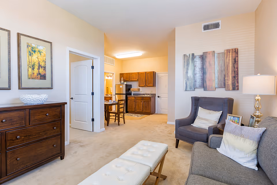 Bright furnished living room with a sofa and armchair, decorative wall art, and a small kitchenette visible in the background.