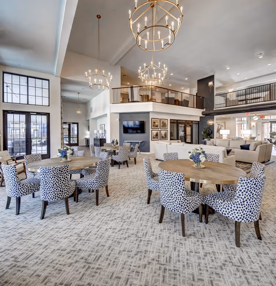 Spacious and well-lit common area with round wooden tables surrounded by blue and white patterned chairs. The room features large windows and glass doors letting in natural light, modern chandeliers hanging from a high ceiling, a seating area with white sofas, and a mezzanine level with additional seating.