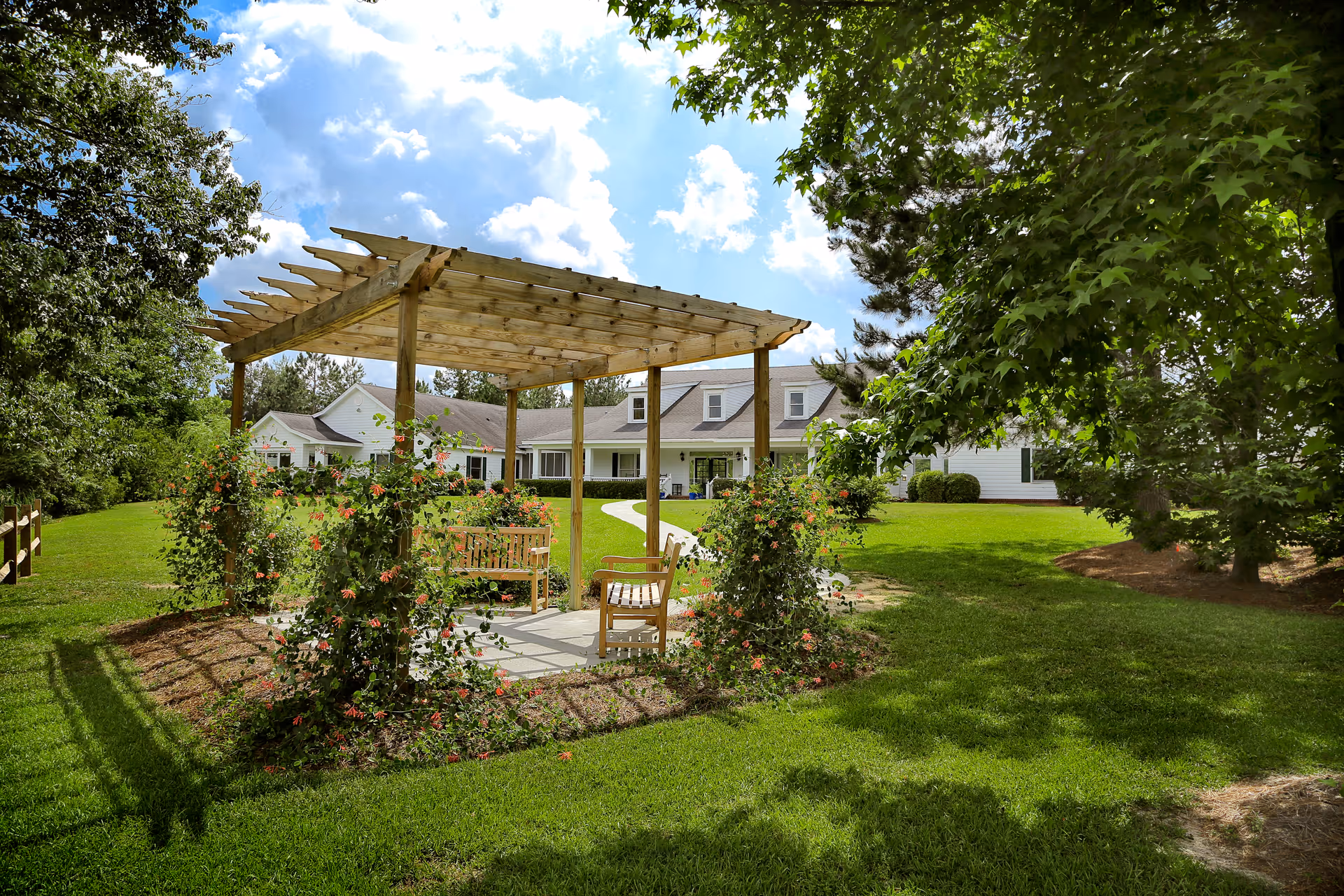 Wooden pergola with benches and flowering vines on a green lawn in front of a senior care building under a partly cloudy sky.
