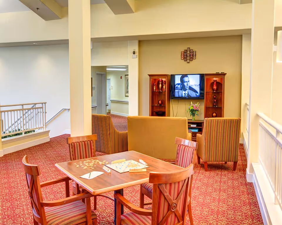 A cozy common area in a senior living facility featuring a wooden table with four striped chairs and a Scrabble game set up on the table. In the background, there are two striped armchairs and a beige loveseat facing a wall-mounted TV displaying a black and white image. The TV is flanked by two wooden shelves with decorative vases. Above the TV, a wall decoration spells out the words LOVE, LAUGH, HOPE, and HELP. The floor is covered with a red patterned carpet, and the area is well-lit with natural and artificial light.