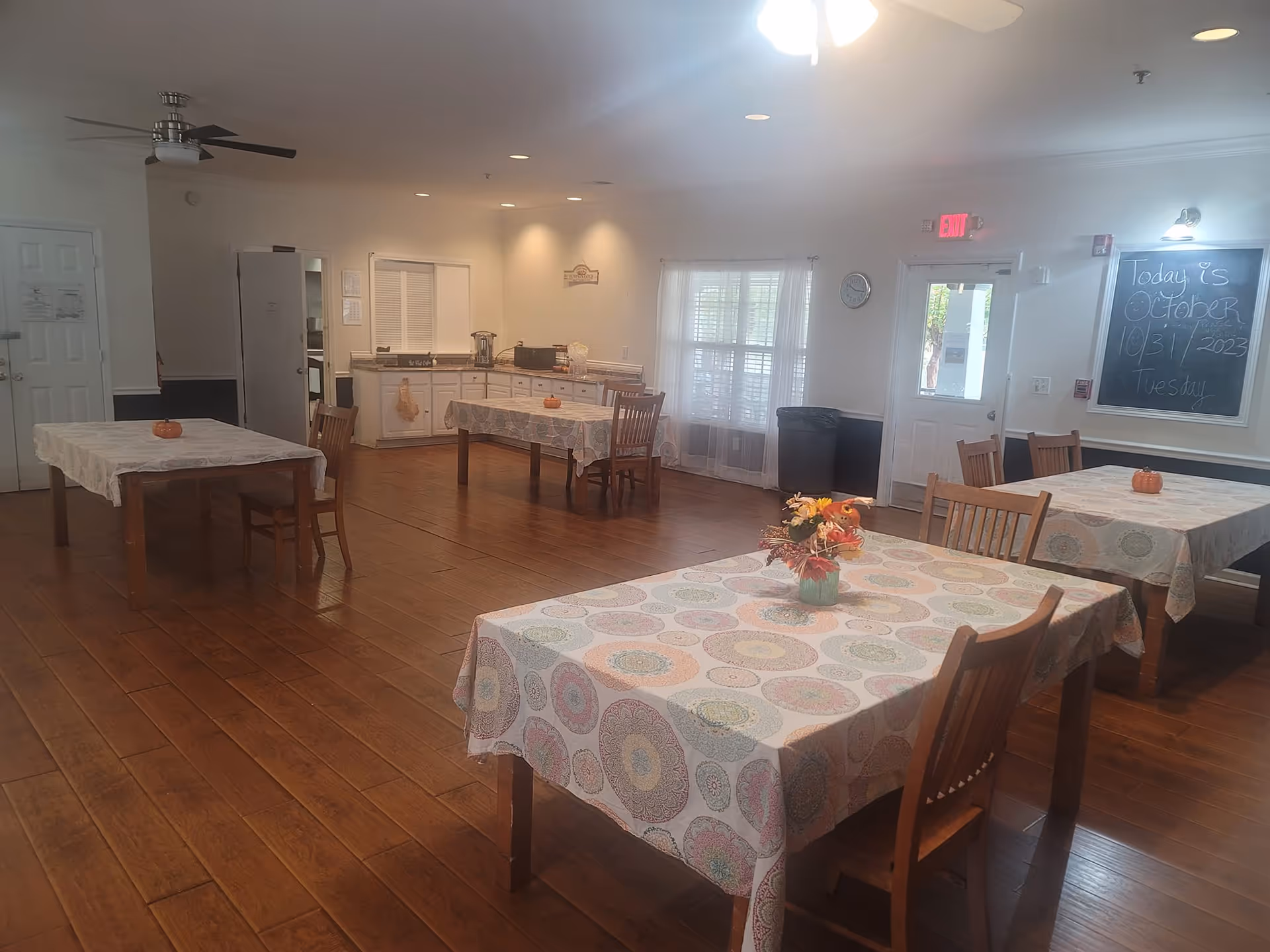 Spacious dining room with several tables covered in patterned tablecloths, wooden chairs, hardwood floors, and a kitchenette area in the back.