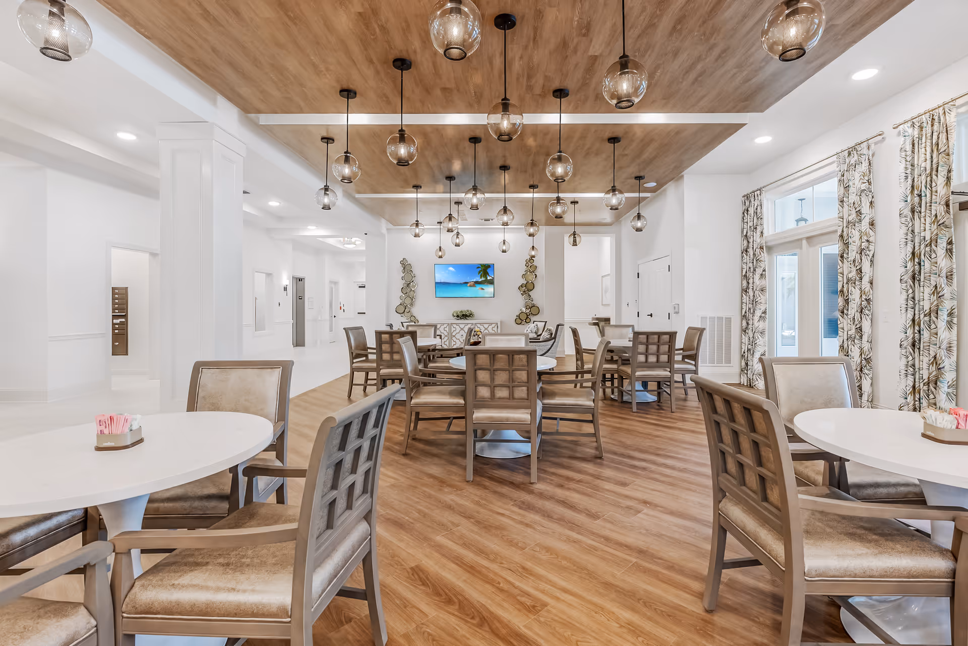 A bright and spacious dining room with multiple round tables and cushioned chairs arranged neatly. The room features a wooden floor and a decorative wooden ceiling panel with hanging globe pendant lights. Large windows with patterned curtains allow natural light to enter. A flat-screen TV is mounted on the far wall above a sideboard, flanked by decorative wall art.