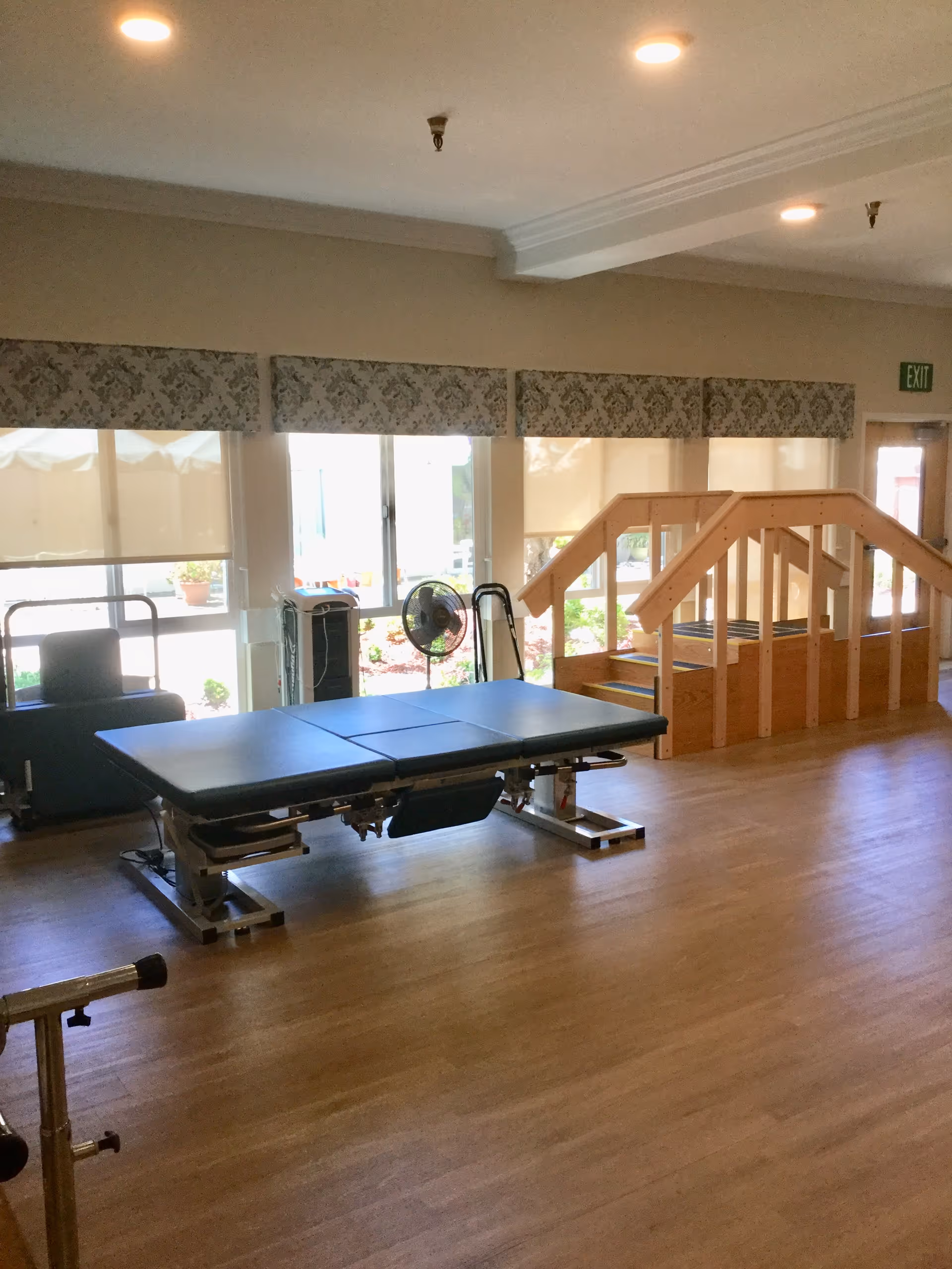 A therapy room with a padded treatment table, a wooden ramp with handrails, large windows with floral valances, and a standing fan. The room has wooden flooring and ceiling lights.
