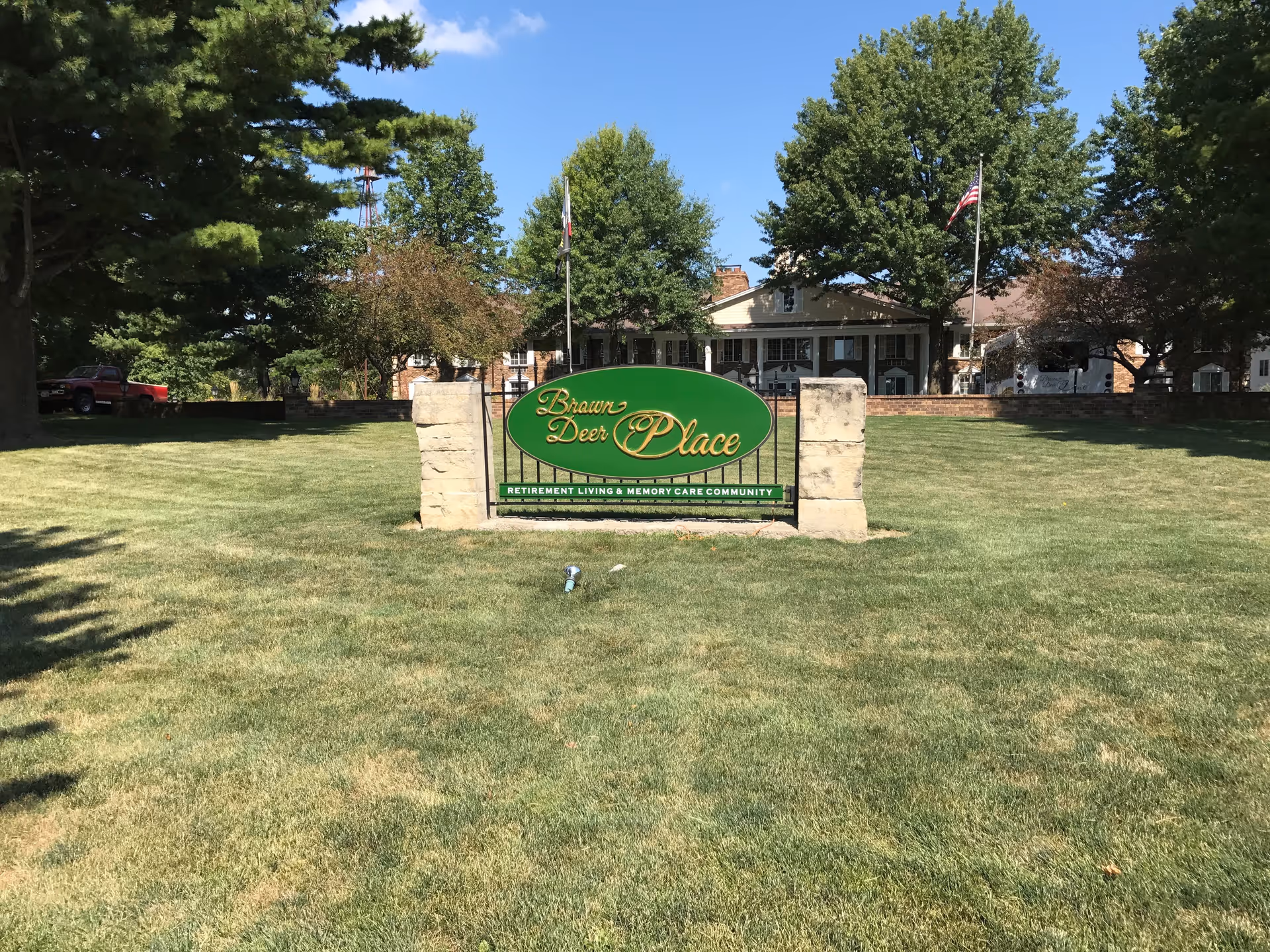 A green sign with gold lettering reading 'Brown Deer Place Retirement Living & Memory Care Community' is displayed on a grassy lawn with trees and a building in the background under a clear blue sky.