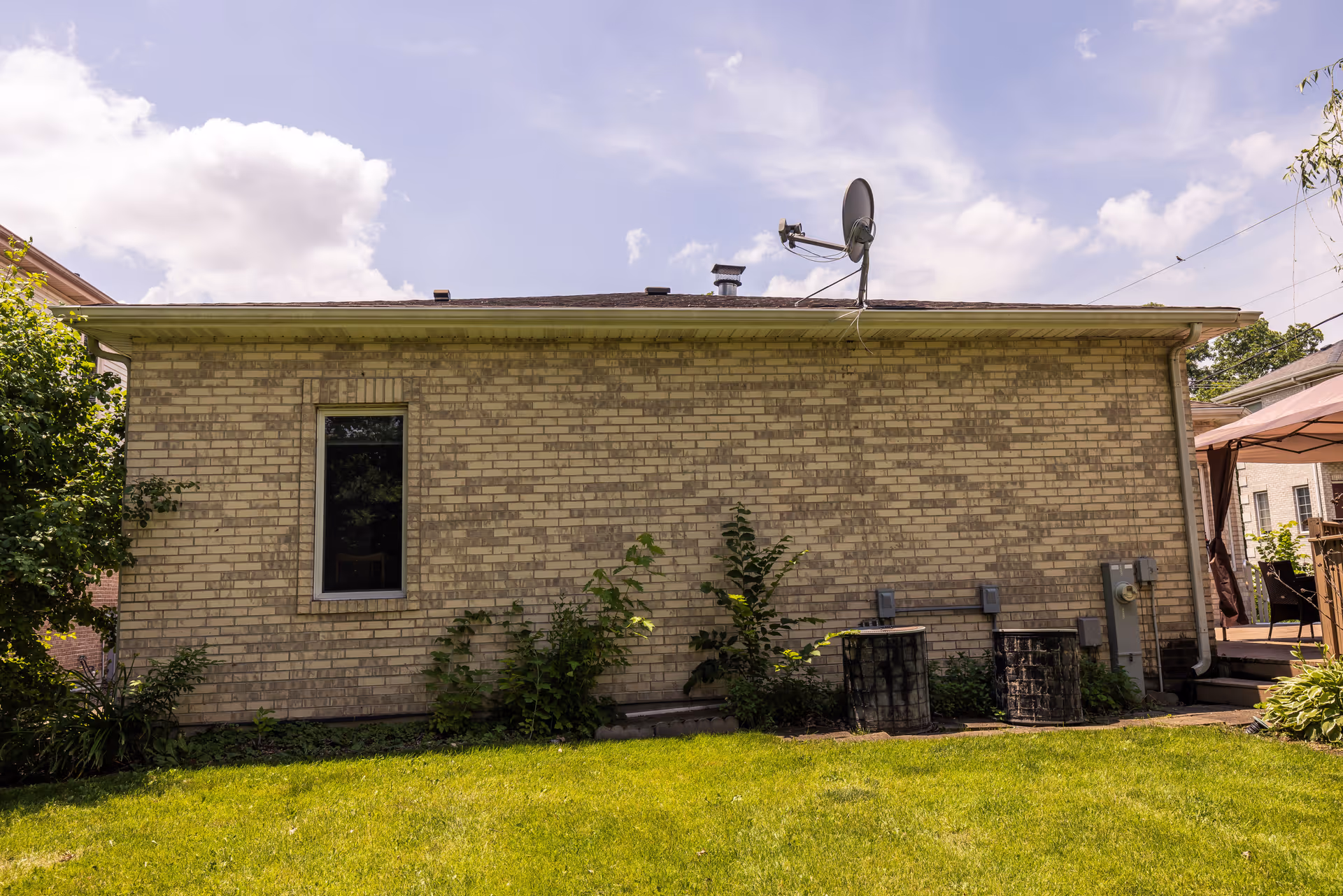 Side exterior view of a single-story brick building with a small window, satellite dish on the roof, two air conditioning units, and some plants growing along the base. There is a green lawn in the foreground and a patio area with outdoor furniture partially visible on the right.