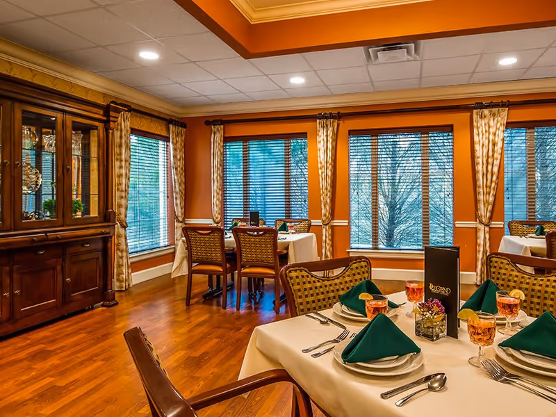 A warmly lit dining room with wooden floors and orange walls. Several tables are set with white tablecloths, green folded napkins, plates, silverware, and glasses filled with a pink beverage. The room has large windows with blinds and patterned curtains, and a wooden cabinet with glass doors displaying glassware.