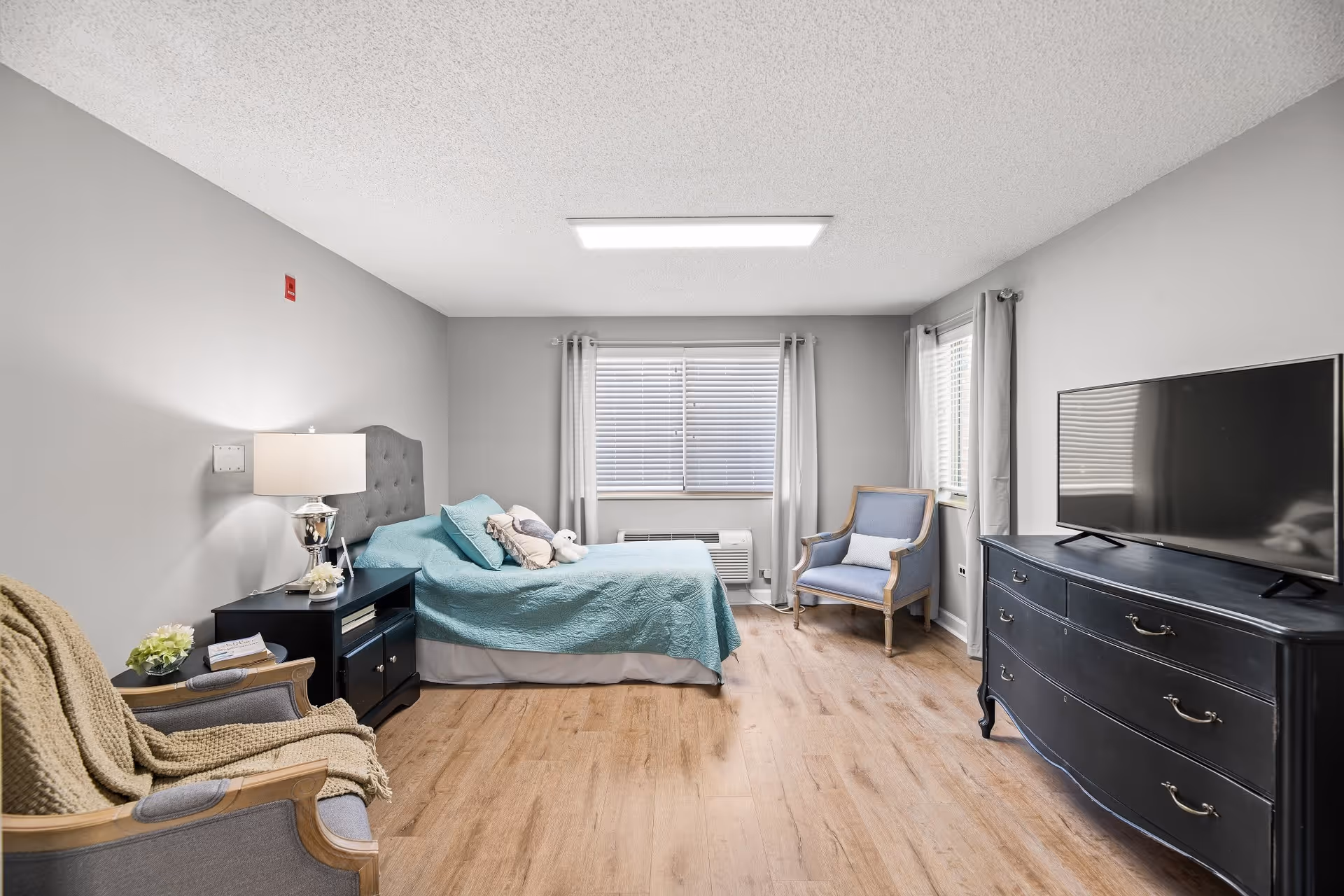 A bright and cozy bedroom with light gray walls and wood flooring. The room features a bed with a teal quilt and decorative pillows, a black nightstand with a lamp, two armchairs with cushions and a throw blanket, a large window with white blinds and curtains, and a black dresser with a flat-screen TV on top.