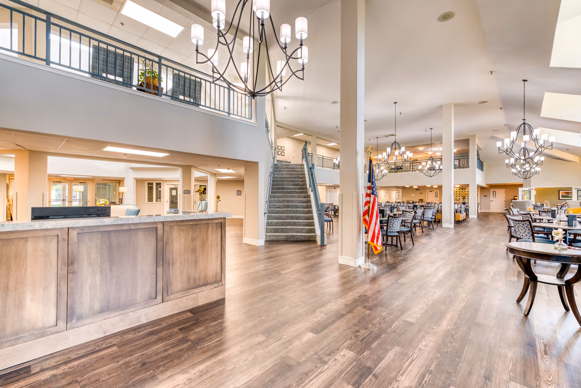 Spacious and well-lit interior of a senior living facility featuring a wooden reception desk on the left, a staircase leading to an upper level in the center, and a large dining area with multiple tables and chairs on the right. The space has high ceilings with modern chandeliers and an American flag near the staircase.