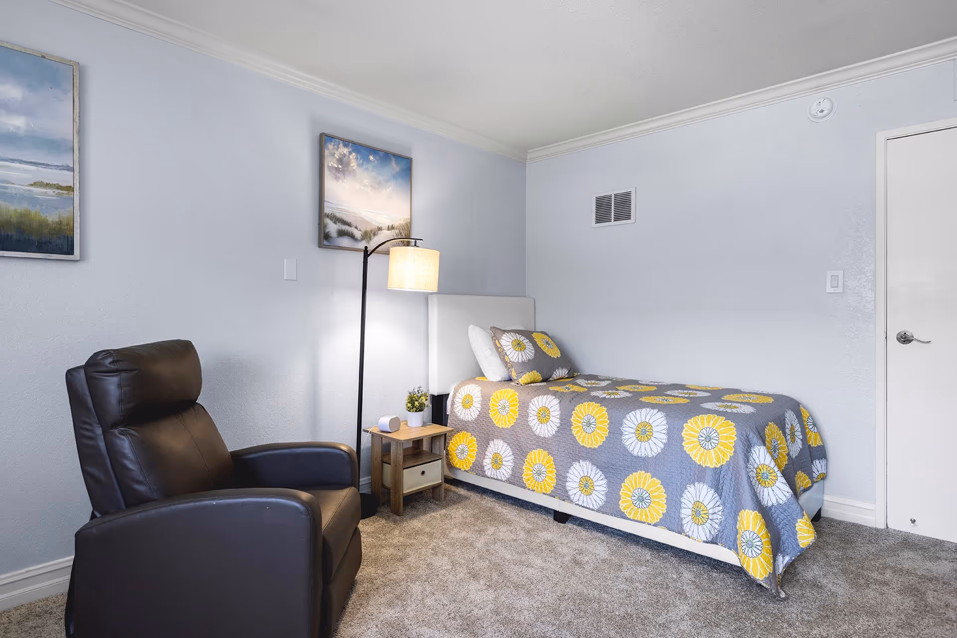 A small bedroom with a single bed covered in a gray quilt with yellow and white flower patterns. Next to the bed is a wooden nightstand with a small plant and a lamp with a beige shade. A black leather recliner chair is positioned near the nightstand. The walls are light gray with two framed landscape paintings hanging above the bed and chair. The floor is carpeted in a neutral tone.