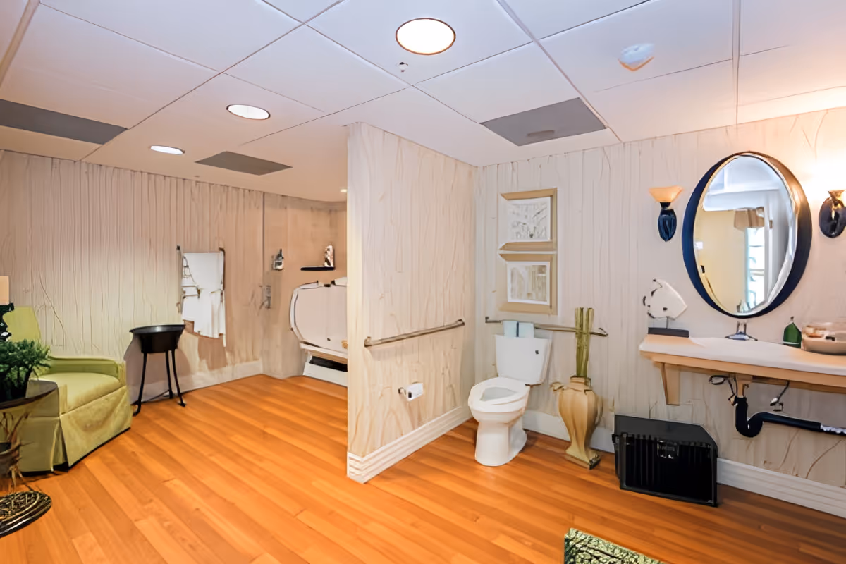Spacious bathroom with wooden flooring featuring a toilet with grab bars, a wall-mounted sink with an oval mirror above it, a walk-in bathtub with a shower, a green armchair, and decorative elements including framed pictures and a vase.