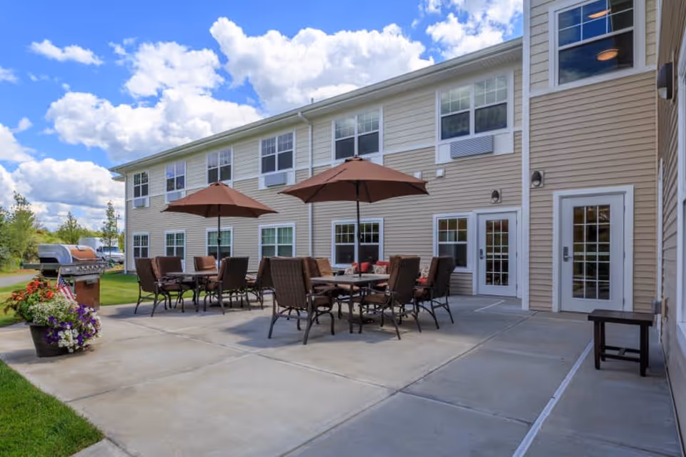 Outdoor patio area at All American Assisted Living at Washington Township featuring multiple tables with brown umbrellas and chairs, a barbecue grill, potted flowers, and the exterior of a beige two-story building with multiple windows and doors under a partly cloudy blue sky.