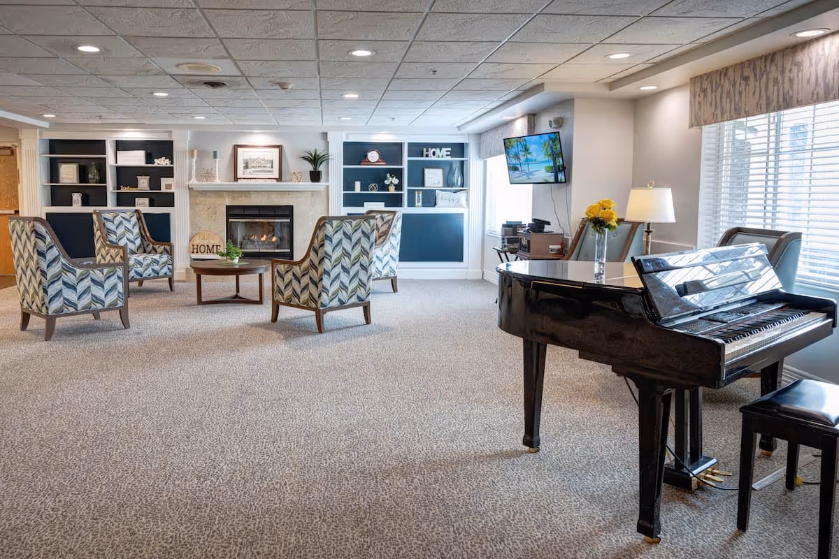 A spacious and well-lit living room area with a patterned carpet, four upholstered armchairs arranged around a round wooden coffee table in front of a fireplace with built-in shelving on either side. A black grand piano with a matching bench is positioned near large windows with blinds. A flat-screen TV is mounted on the wall, and a table lamp and vase with yellow flowers sit on a small table next to the piano.