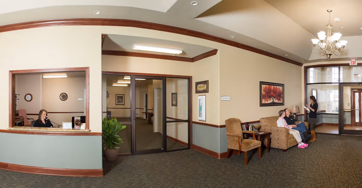 A senior living facility reception area with a receptionist sitting behind a counter on the left, a hallway with glass doors in the center, and a seating area on the right where two elderly women are sitting on a couch and a younger woman is standing and talking to them. The room has beige walls with wooden trim, carpeted floor, a chandelier, and framed artwork on the wall.