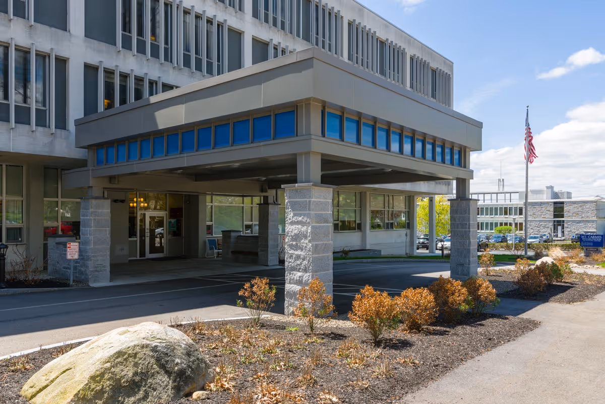 Entrance area of Mt. Carmel Rehabilitation and Nursing Center showing a covered drop-off zone supported by stone pillars, a landscaped area with small bushes and a large rock, and an American flag on a flagpole. The building has multiple windows and a clear blue sky is visible.