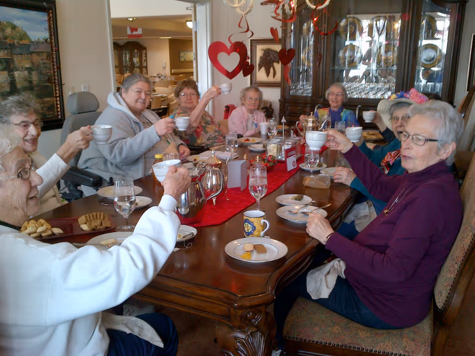 A group of elderly women sitting around a wooden dining table raising teacups in a toast. The table is set with plates, cups, glasses, and a red table runner. The room has a china cabinet with glassware and plates, and heart-shaped decorations hanging from the ceiling.