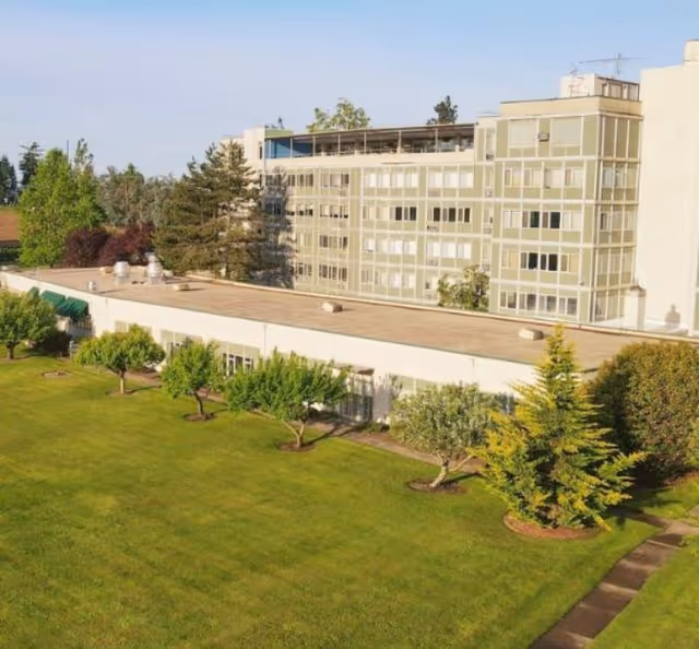 Exterior view of a multi-story building with large windows, surrounded by a well-maintained green lawn and several trees under a clear sky.