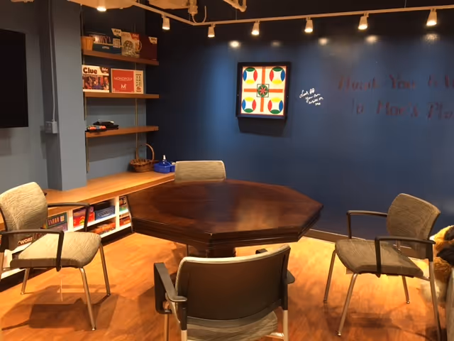 Activity/common room with an octagonal wooden table surrounded by chairs, shelves of board games, and a blue accent wall with artwork.
