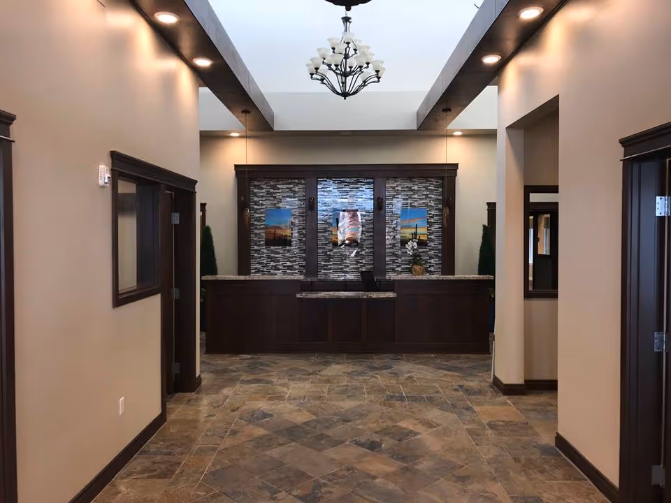 Spacious lobby with a central dark wood reception desk, tiled floor, chandelier, and decorative stone wall behind the desk.