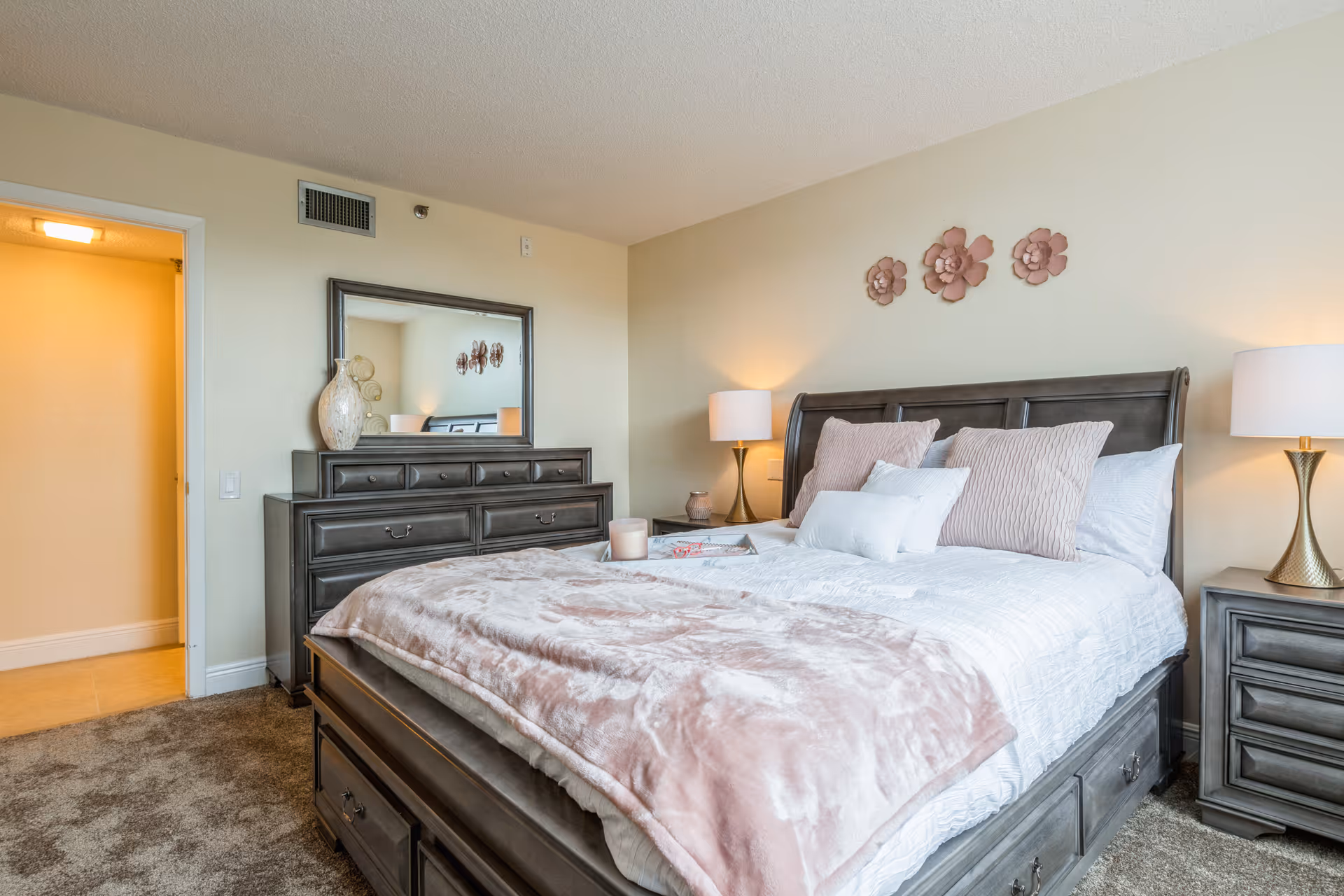A cozy bedroom with a large bed featuring white and light pink bedding and pillows. There are two bedside tables with matching lamps on either side of the bed. A dresser with a large mirror and decorative vase is positioned against the wall. Three pink flower wall decorations hang above the bed. The room has beige walls and carpeted flooring, with an open doorway leading to another room.