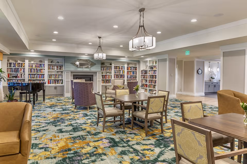 A spacious and well-lit common area with patterned carpet, several tables and chairs, armchairs, a grand piano, and built-in bookshelves filled with books surrounding a fireplace with a decorative mirror above it.