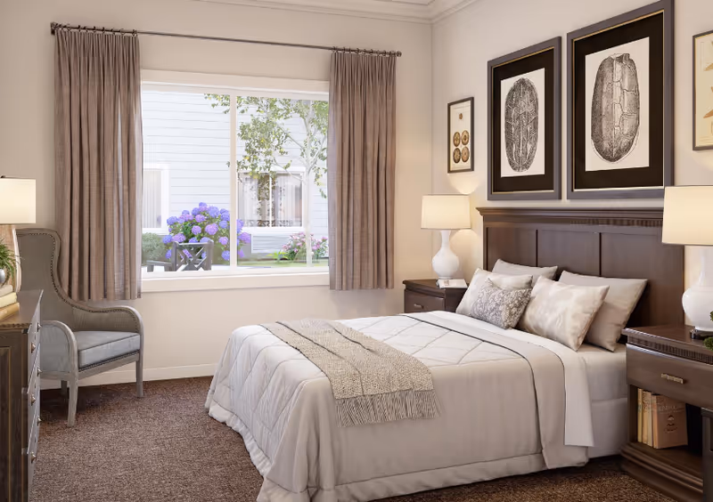 A cozy bedroom with a neatly made bed featuring white and beige bedding and multiple pillows. On either side of the bed are dark wooden nightstands with white lamps. Above the bed are three framed botanical prints. A large window with beige curtains reveals a view of a garden with purple flowers and greenery. A gray upholstered chair is positioned near a dresser with books and a small plant.