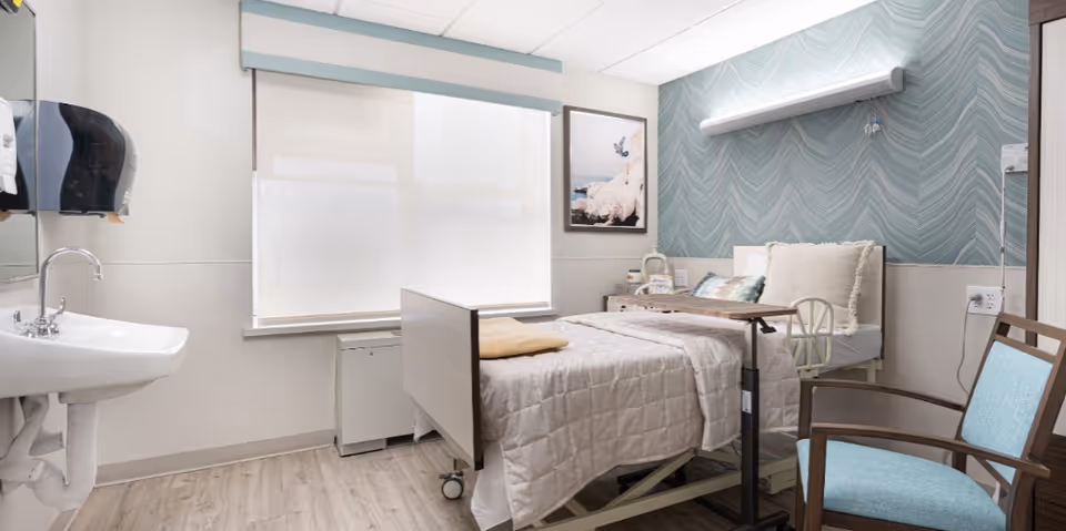 A bright and clean healthcare facility bedroom with a hospital bed covered in a beige quilt, a wooden overbed table, a blue cushioned chair, a wall-mounted light, a sink with a soap dispenser, and a large window with a white shade. The wall behind the bed has a blue patterned wallpaper and a framed picture.
