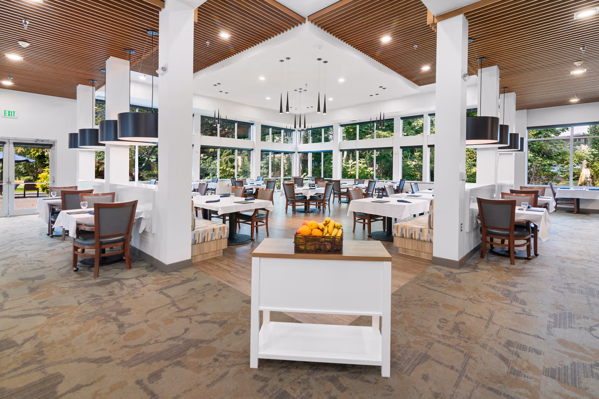 Bright, spacious dining room with tables and chairs, large windows and pendant lights, and a small cart topped with a basket of fruit in the foreground.