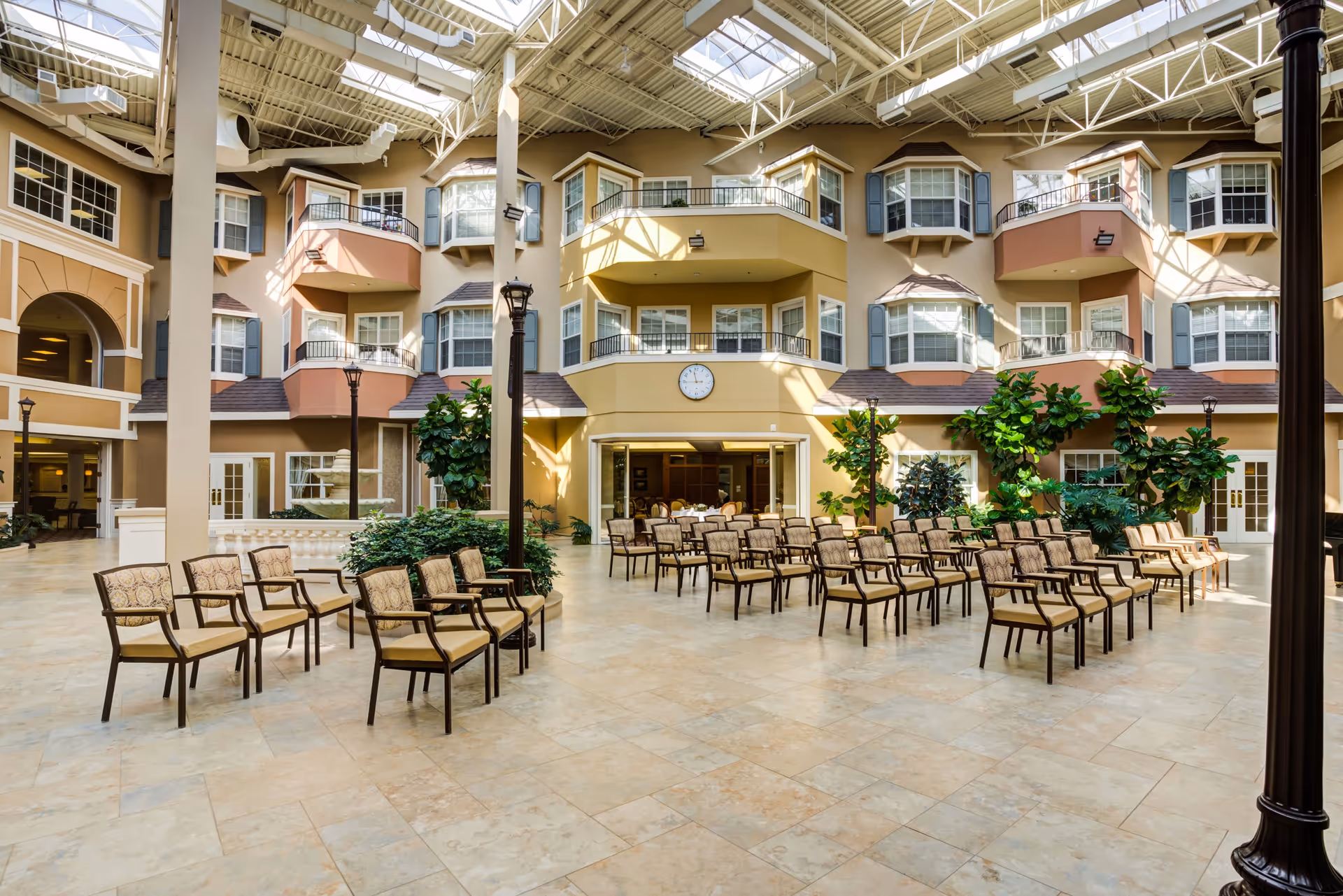 Spacious sunlit indoor atrium with rows of chairs, potted plants, surrounding balconies, and large skylights.