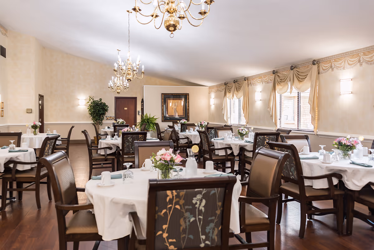 A well-lit dining room in a senior living facility with multiple round tables covered with white tablecloths. Each table is set with cups, glasses, napkins, and small floral centerpieces. The room features wooden chairs with patterned upholstery, chandeliers hanging from the ceiling, wall sconces, and windows with draped curtains allowing natural light to enter.