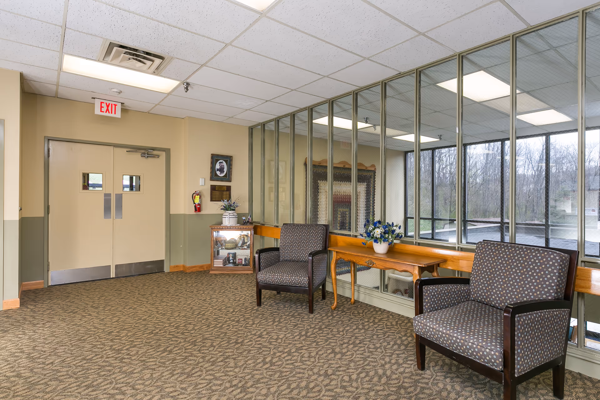 A waiting area inside a facility with two patterned armchairs, a wooden table with a vase of flowers, a glass display cabinet, and large windows showing an outdoor view of trees. The walls are painted beige and green, and there is a double door with an exit sign above it.