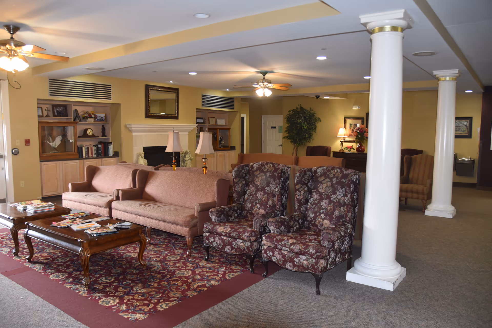A cozy assisted living facility common area with beige walls, two floral-patterned armchairs, two pink sofas, wooden coffee tables with magazines, a fireplace with a mirror above it, ceiling fans with lights, decorative columns, and various lamps and plants providing a warm ambiance.
