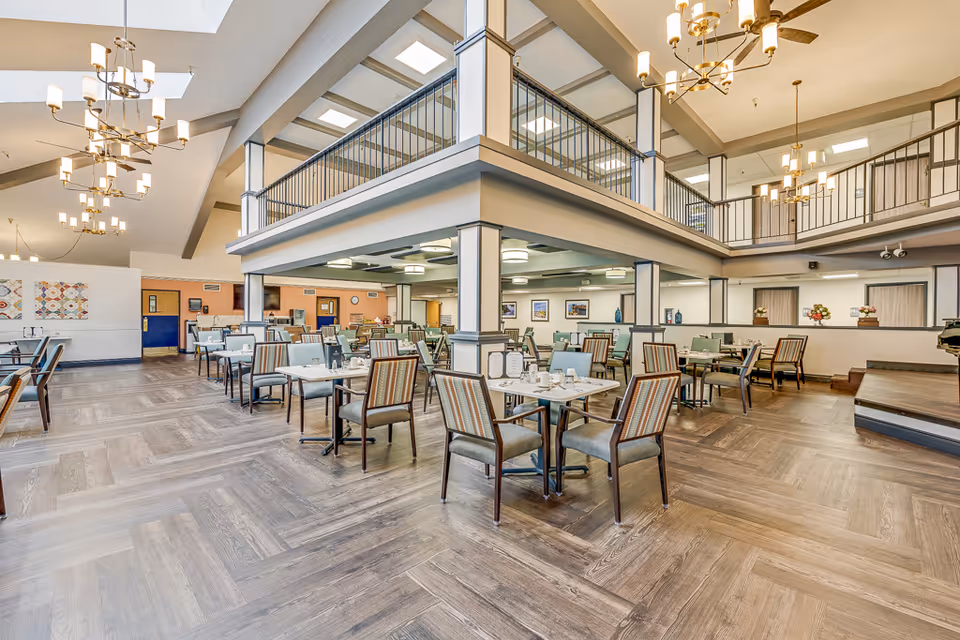 Spacious double-height dining room with multiple tables and chairs, chandeliers, and a mezzanine balcony in a senior living facility.
