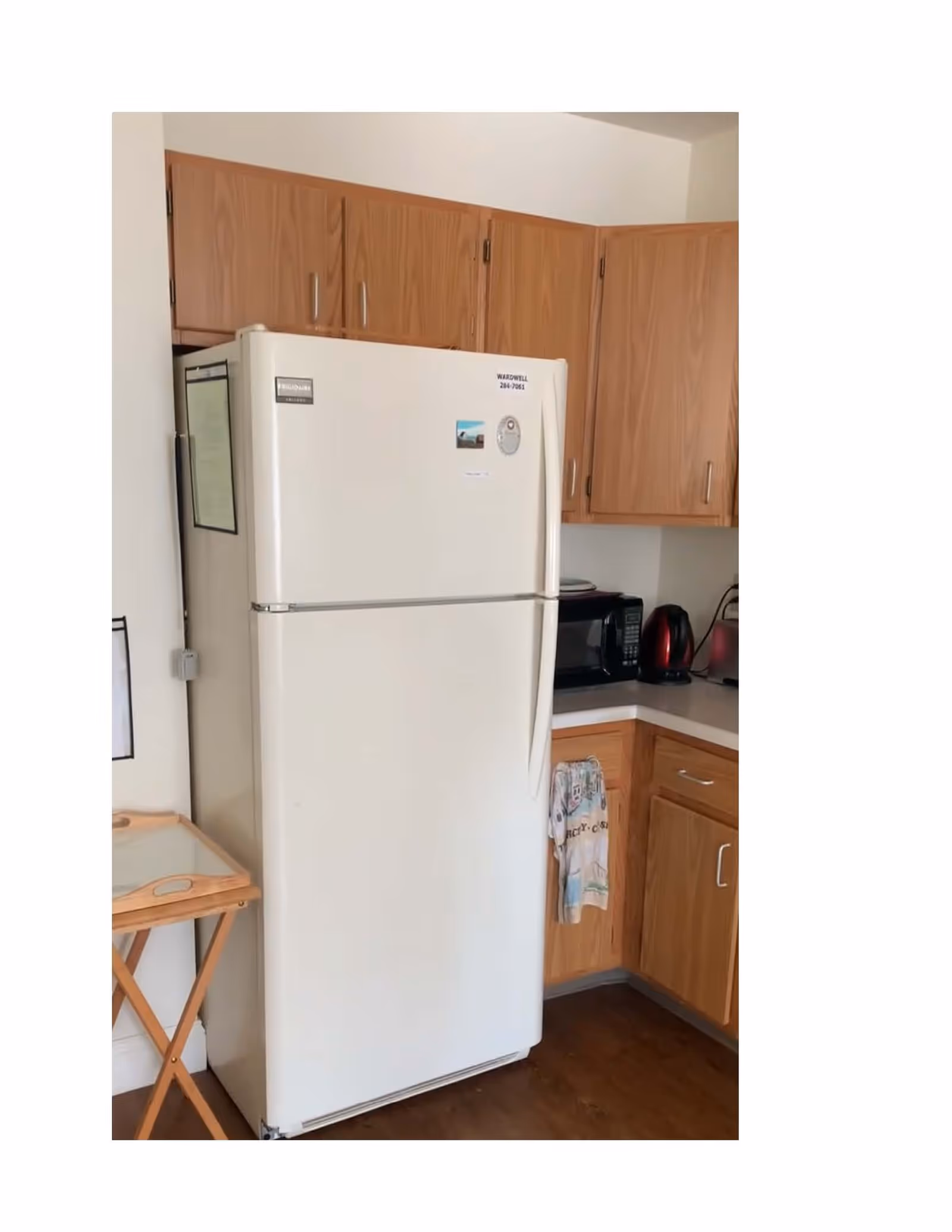 A kitchen corner featuring a white refrigerator with wooden cabinets above and beside it. On the countertop next to the refrigerator, there is a microwave, a red electric kettle, and a toaster. A dish towel hangs from a cabinet handle, and a small wooden tray table is positioned to the left of the refrigerator.