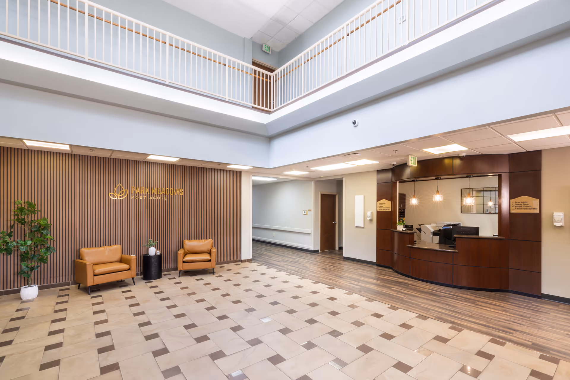 Spacious and well-lit lobby area of Park Meadows Post Acute facility featuring a tiled floor with a checkered pattern, two tan leather chairs with a small black round table and plants against a wooden slat wall with the facility's gold logo. To the right is a curved wooden reception desk with pendant lights above and a hallway leading further into the building.