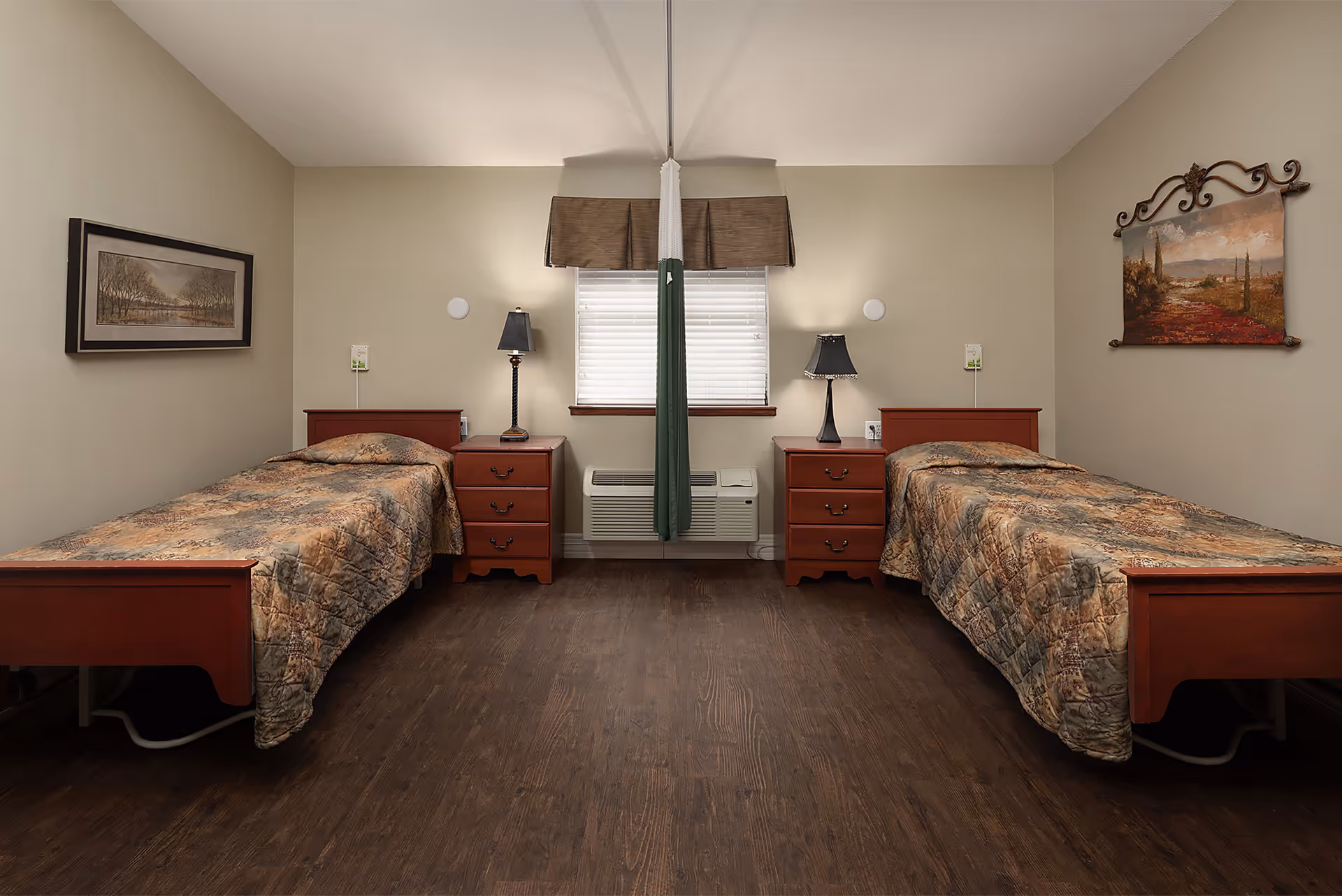 A simple, clean bedroom with two single beds separated by a window with blinds and a brown valance. Each bed has a wooden headboard and footboard, a matching nightstand with a lamp, and a patterned bedspread. There are framed landscape paintings on each side wall and a dark wood floor.