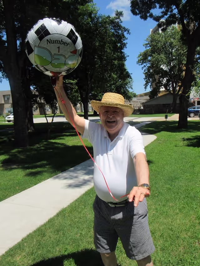 An elderly man wearing a straw hat, white polo shirt, and checkered shorts is standing on a sidewalk in a grassy outdoor area with trees and buildings in the background. He is smiling and holding a balloon that says 'Number 1 Dad'.