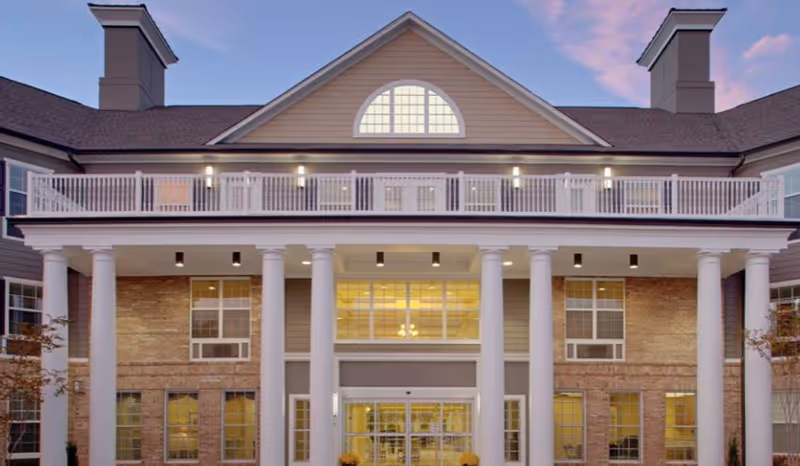 Front facade of a multi-story senior living building with white columns, a second-story balcony, and large illuminated windows.