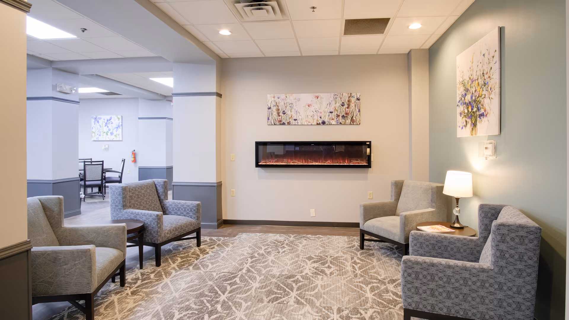 A cozy senior living common area with four upholstered armchairs arranged around a patterned area rug. A modern electric fireplace is mounted on the wall, with floral artwork above it. A small side table with a lamp and a book is placed between two chairs. The room has soft lighting and a calm, neutral color palette with light blue and beige walls.