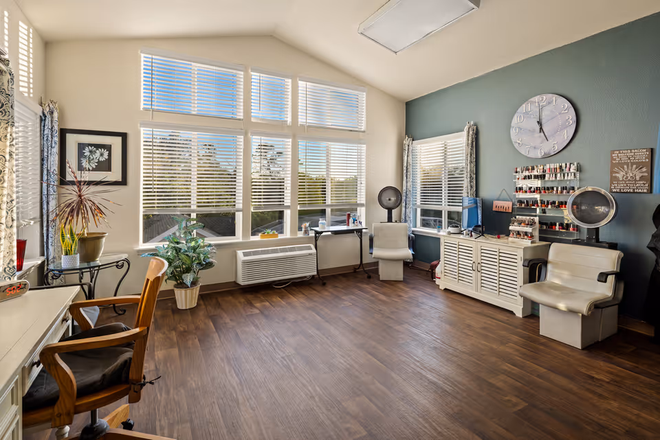 Bright senior living salon room with large windows, seating, nail polish racks, and hair-drying chairs.