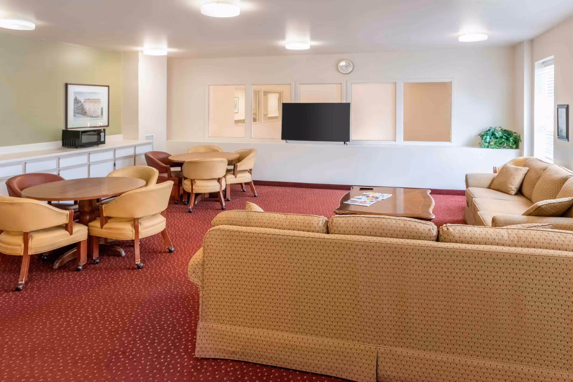 A senior living community common area with beige sofas, a wooden coffee table with magazines, round tables with beige and brown chairs, a wall-mounted flat screen TV, a microwave on a counter, and framed artwork on the walls. The room has red carpet flooring and white walls with windows and a clock.