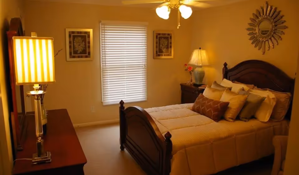 A warmly lit bedroom with a wooden bed frame, neatly made bed with multiple pillows, a nightstand with a lamp and flowers, a dresser with a lamp, two framed pictures on the wall, a window with closed blinds, and a decorative sunburst mirror above the bed.