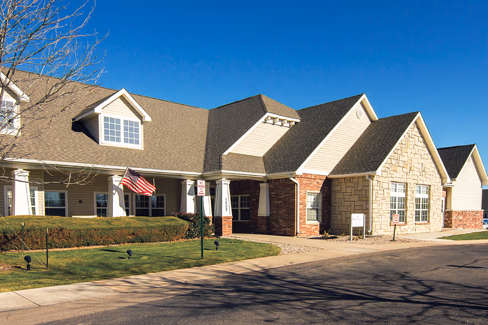 Exterior view of a single-story senior living facility building with a combination of brick, stone, and siding walls, a gabled roof, and an American flag displayed near the entrance under a clear blue sky.